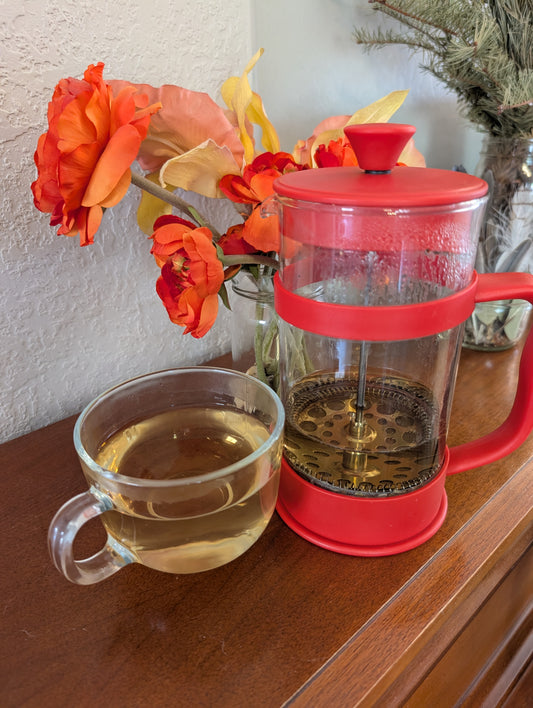Red French press and clear mug with tea on a wooden surface with orange flowers in the background.