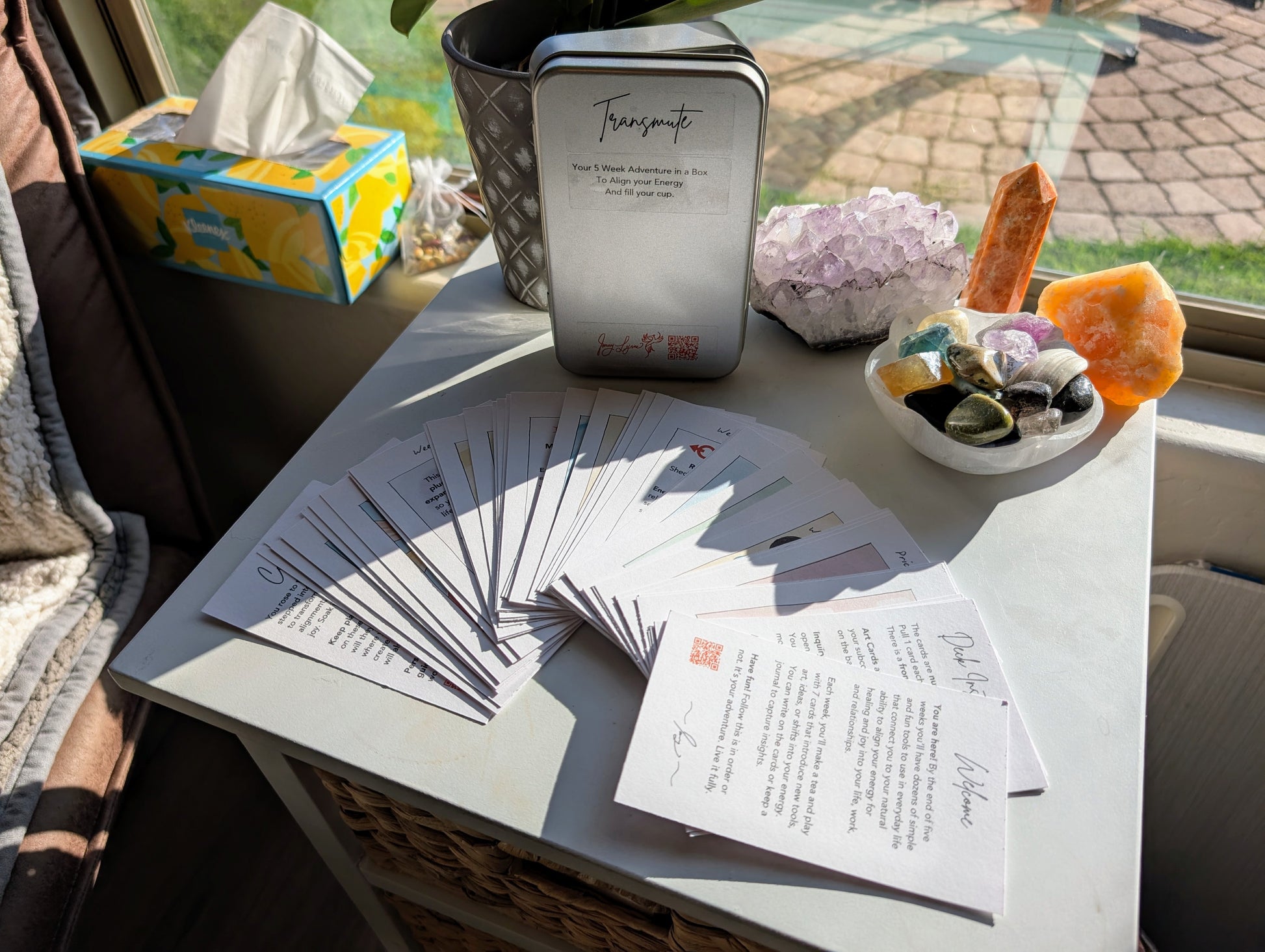 Table with colorful art cards, crystals, and a tin near a sunny window on a white table.